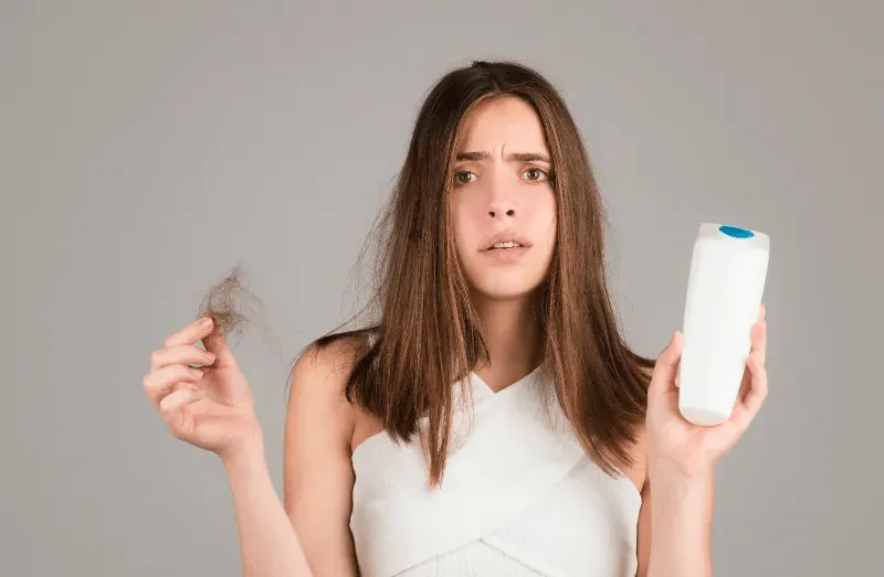 The image depicts a woman holding a bunch of hair in one hand and a shampoo bottle in the other, expressing concern over hair loss and seeking solutions.