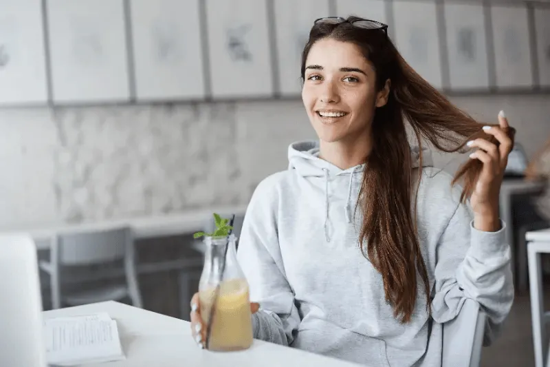 The image shows a woman smiling while holding a drink and playfully touching her hair, symbolizing a relaxed and balanced approach to maintaining healthy hair.