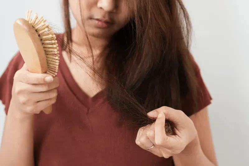 The image shows a woman holding a hairbrush with visible hair strands tangled in it, illustrating concerns about hair loss and the need to understand its underlying causes.