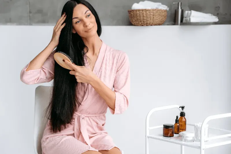 The image shows a woman in a pink robe brushing her long hair, showcasing the importance of consistent hair care.